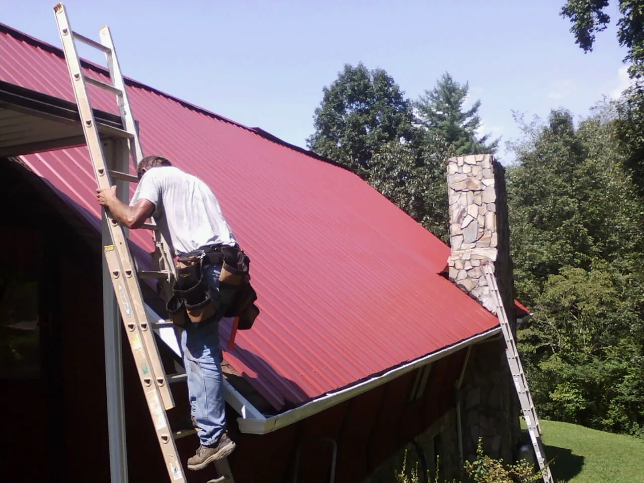 Roofer on a ladder beside a red metal roof and stone chimney — Soffit & Fascia Repair by Roofing Brothers in El Dorado Springs